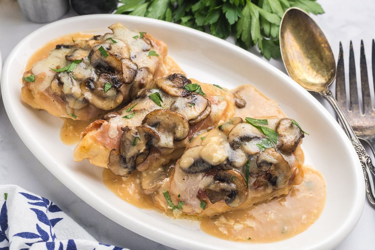 Three chicken breasts prepared chicken Valdostana style, topped with sautéed mushrooms and creamy sauce, garnished with parsley on a white plate.