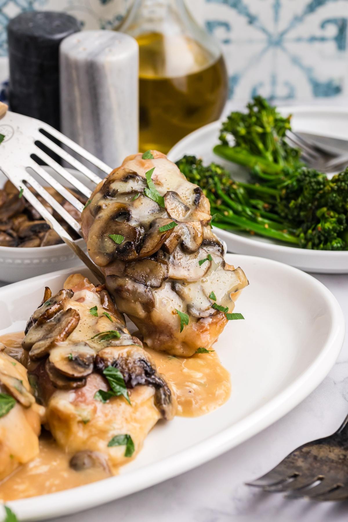 Chicken topped with creamy mushroom sauce on a white plate, with broccolini in the background.