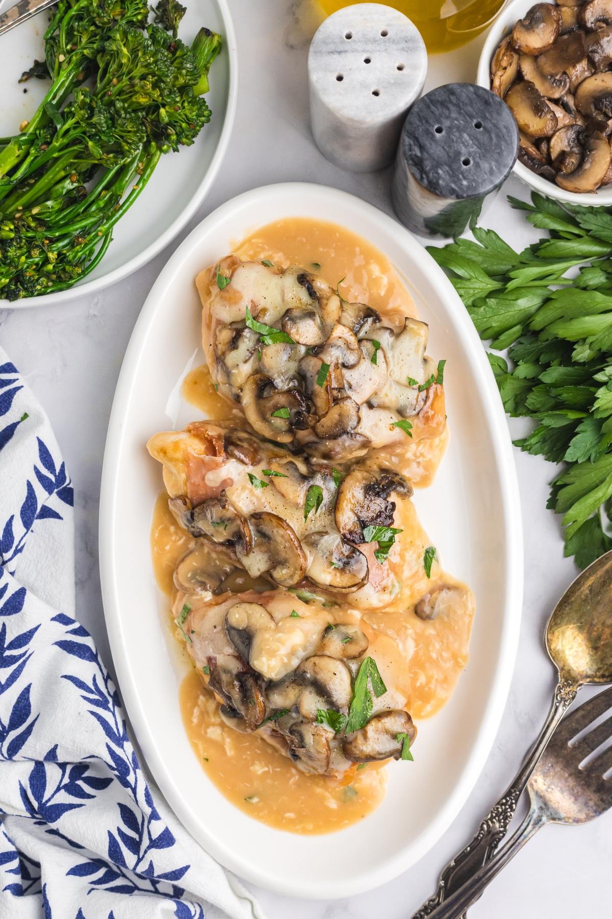 Three chicken breasts with mushroom sauce on a white plate, garnished with parsley, next to broccolini and herbs.