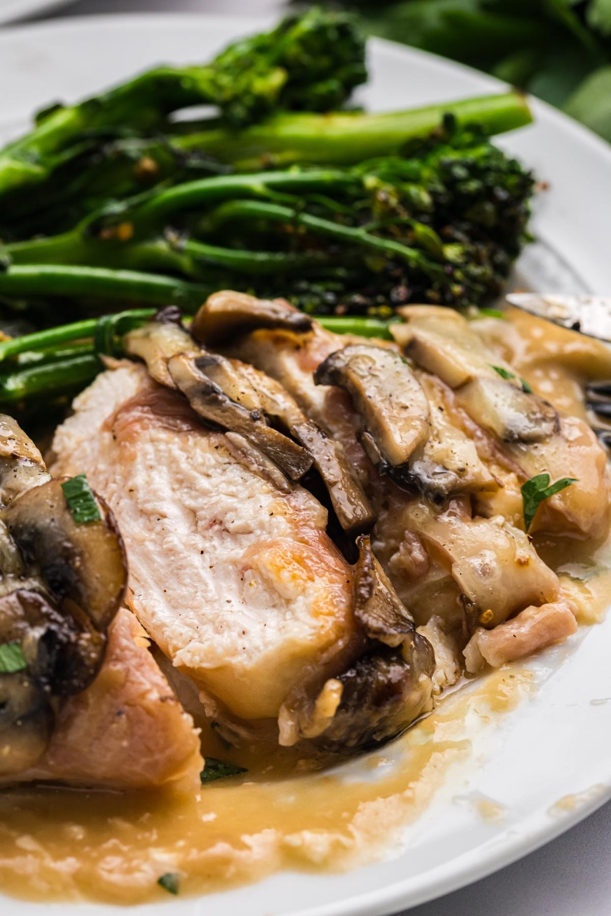 Sliced chicken breast topped with mushrooms in sauce, served with broccolini on a white plate.