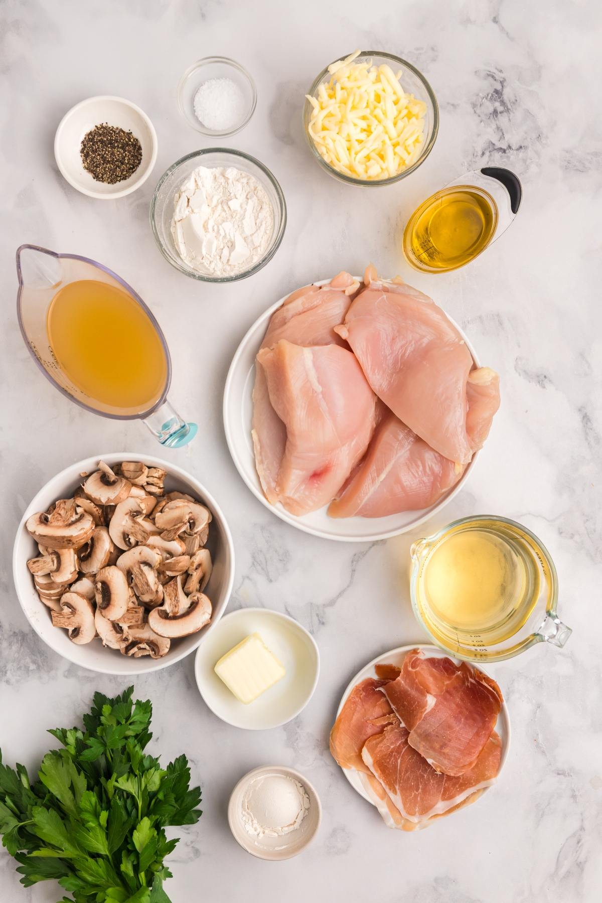 Raw chicken breasts, prosciutto, mushrooms, cheese, broth, wine, flour, butter, oil, spices, and parsley on a table.