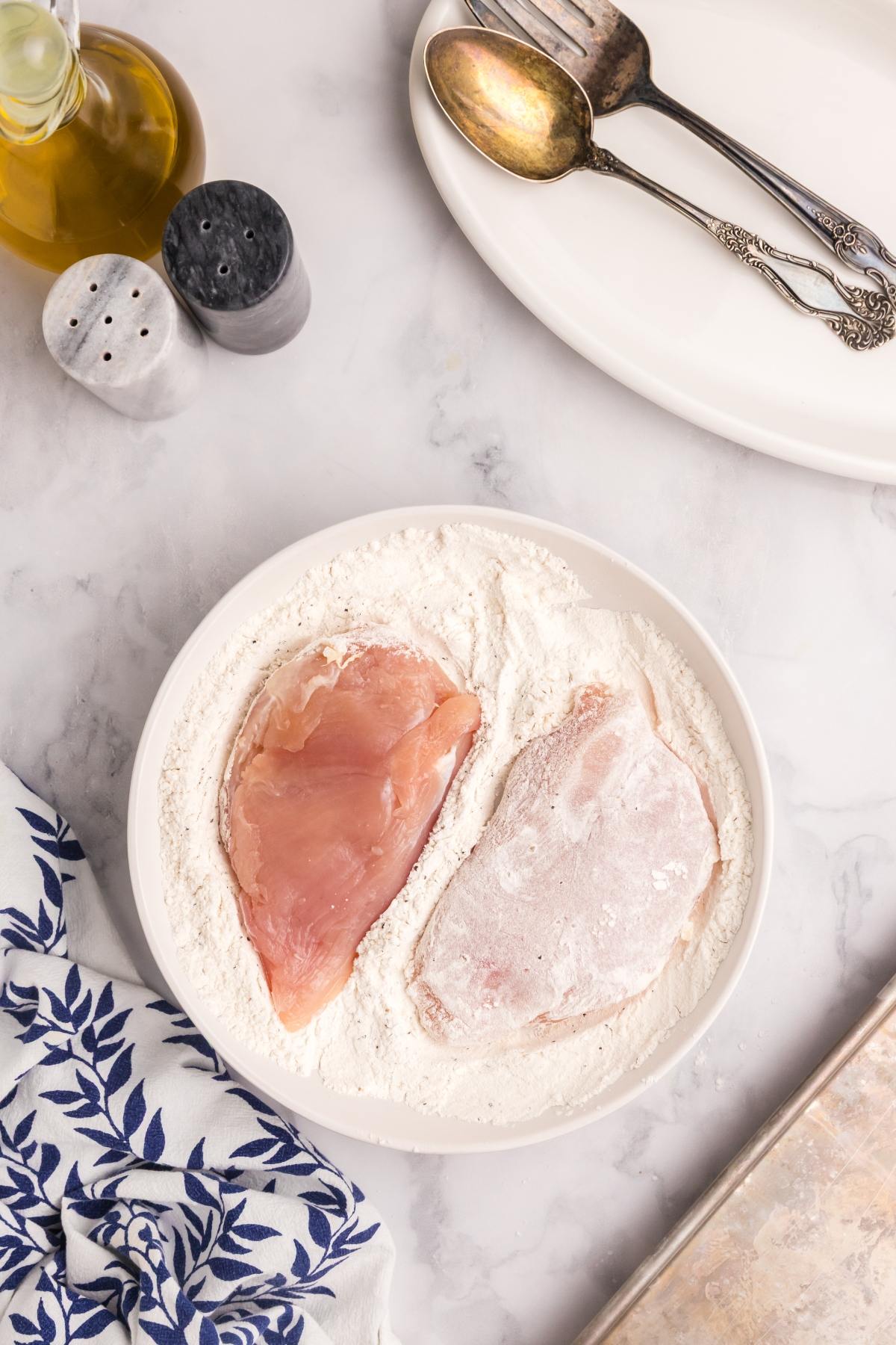 Two raw chicken breasts in a bowl of flour, beside olive oil, salt, and pepper shakers on a marble counter.