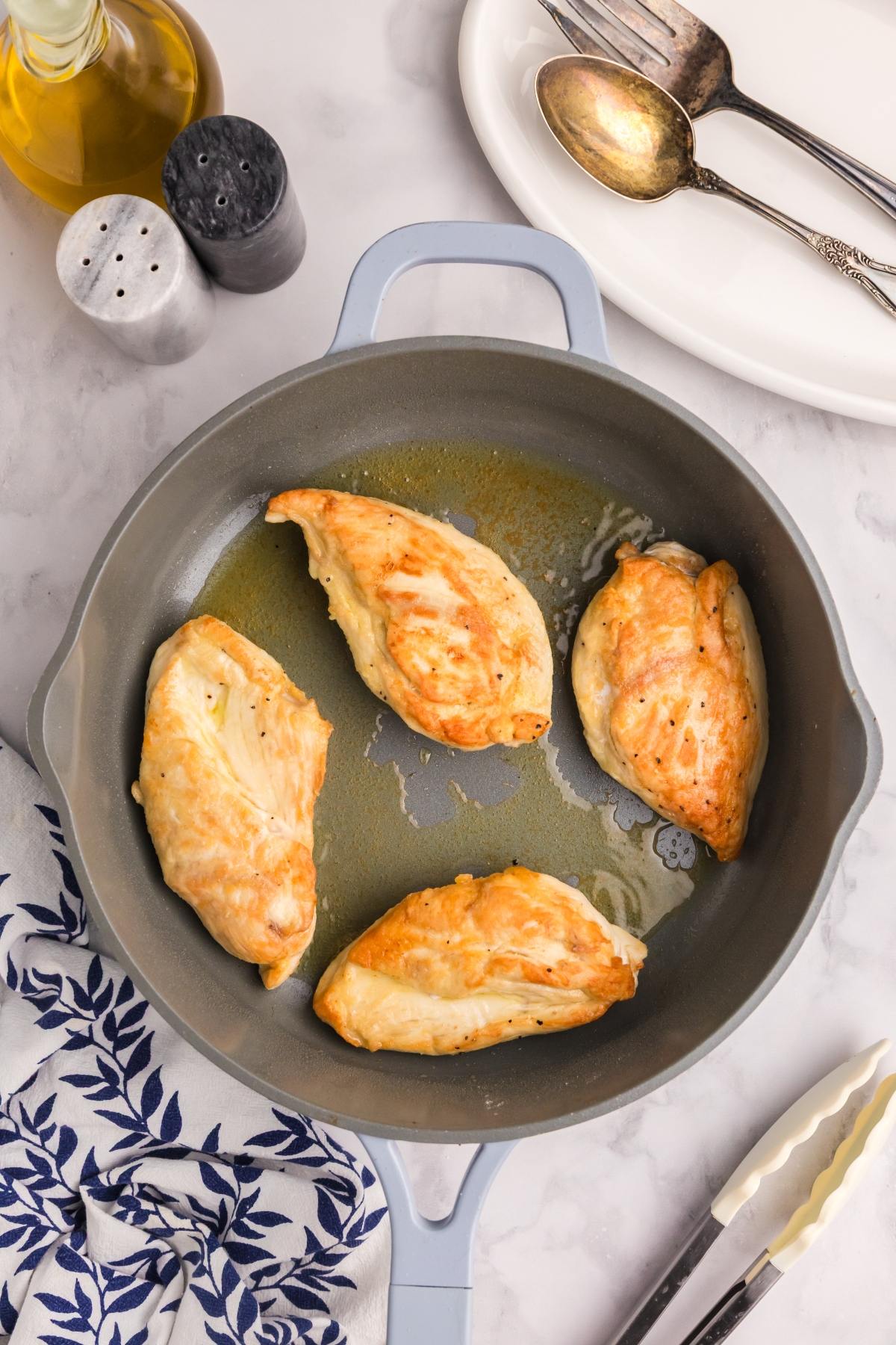 Four cooked chicken breasts in a skillet, with utensils, plates, oil, and seasoning nearby on a marble counter.