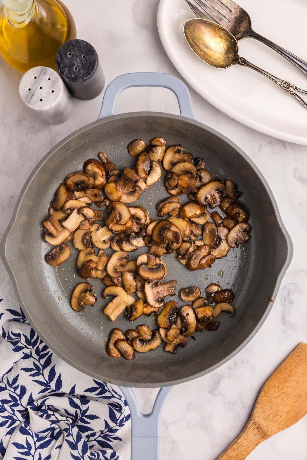 Sliced mushrooms sautéing in a gray skillet, with utensils, plates, and seasonings nearby on a marble surface.