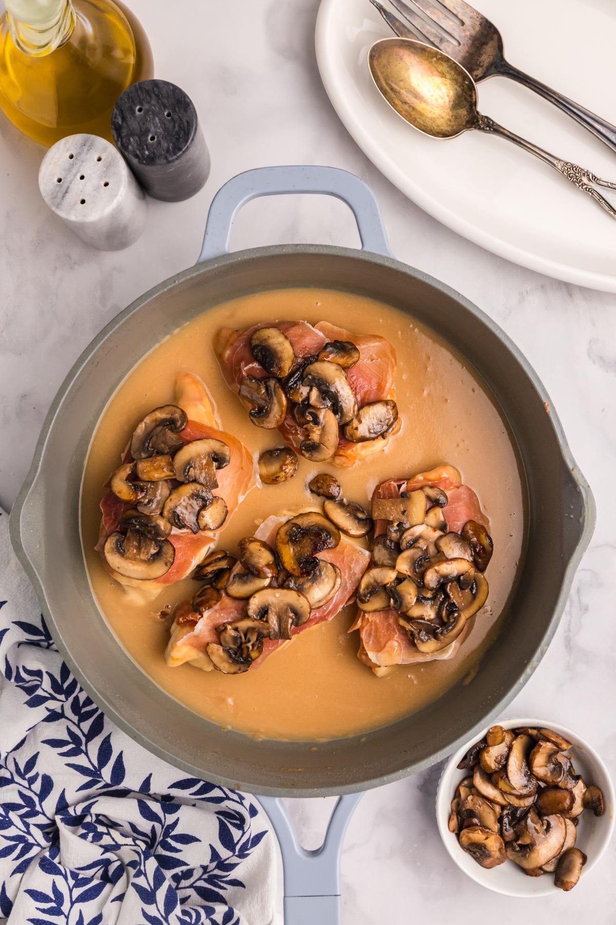 Chicken breasts in a creamy sauce topped with sautéed mushrooms in a pan, next to a bowl of mushrooms.