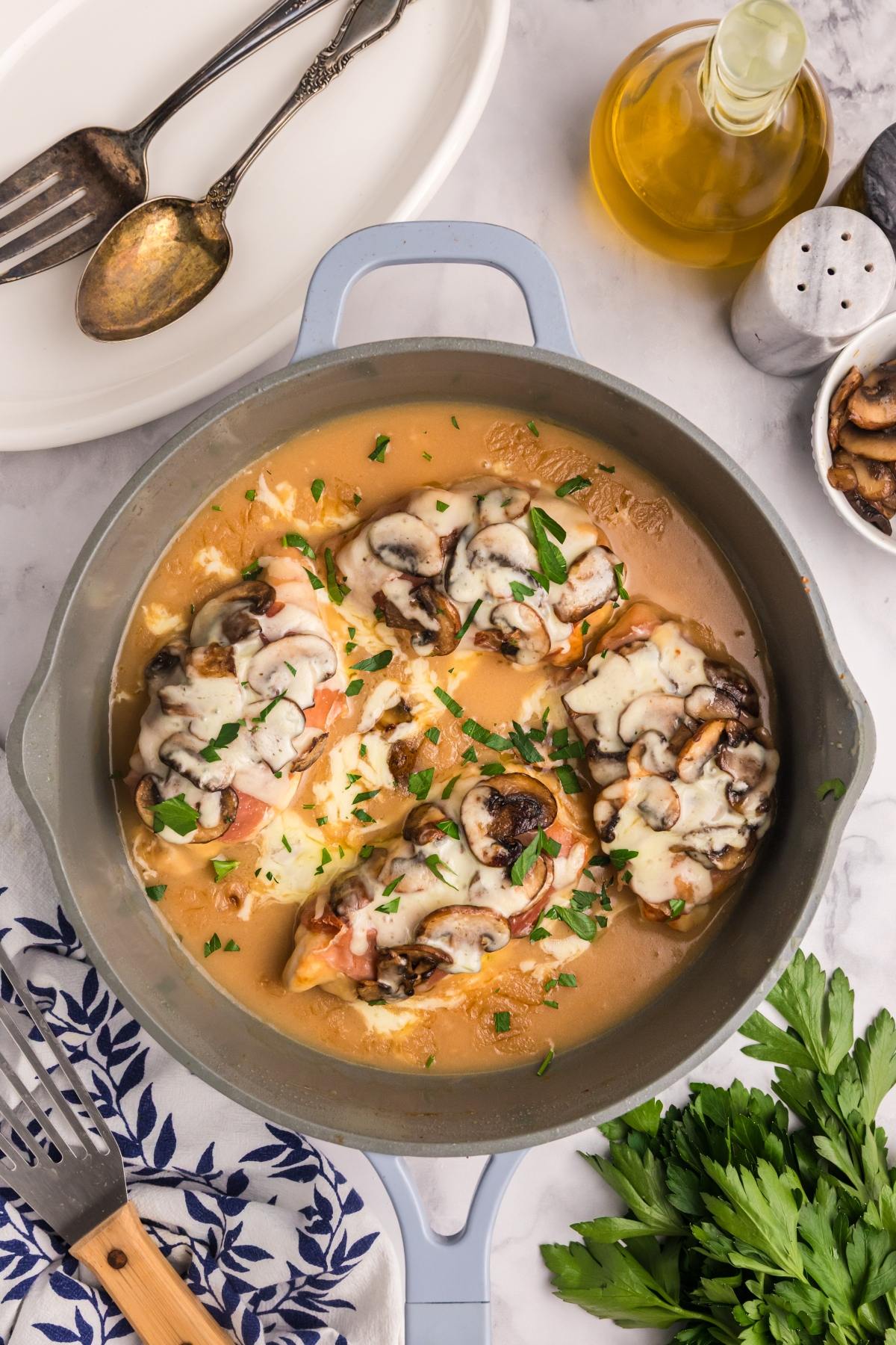 Chicken breasts topped with mushrooms and melted cheese in a creamy sauce, garnished with parsley in a pan.