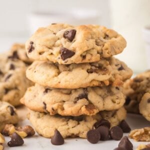 Chocolate chip walnut cookies stacked on a table with scattered chocolate chips and walnuts.