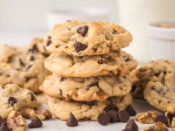 Chocolate chip walnut cookies stacked on a table with scattered chocolate chips and walnuts.