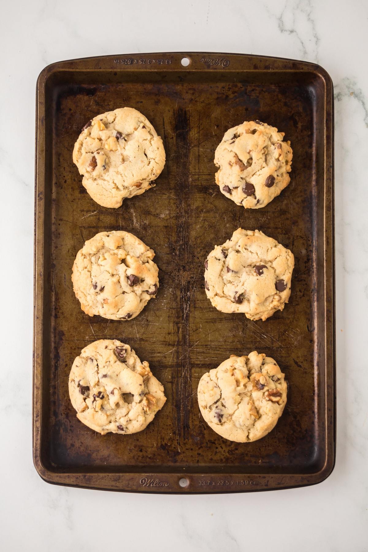Six chocolate chip cookies on a metal baking sheet, placed on a white marble surface.