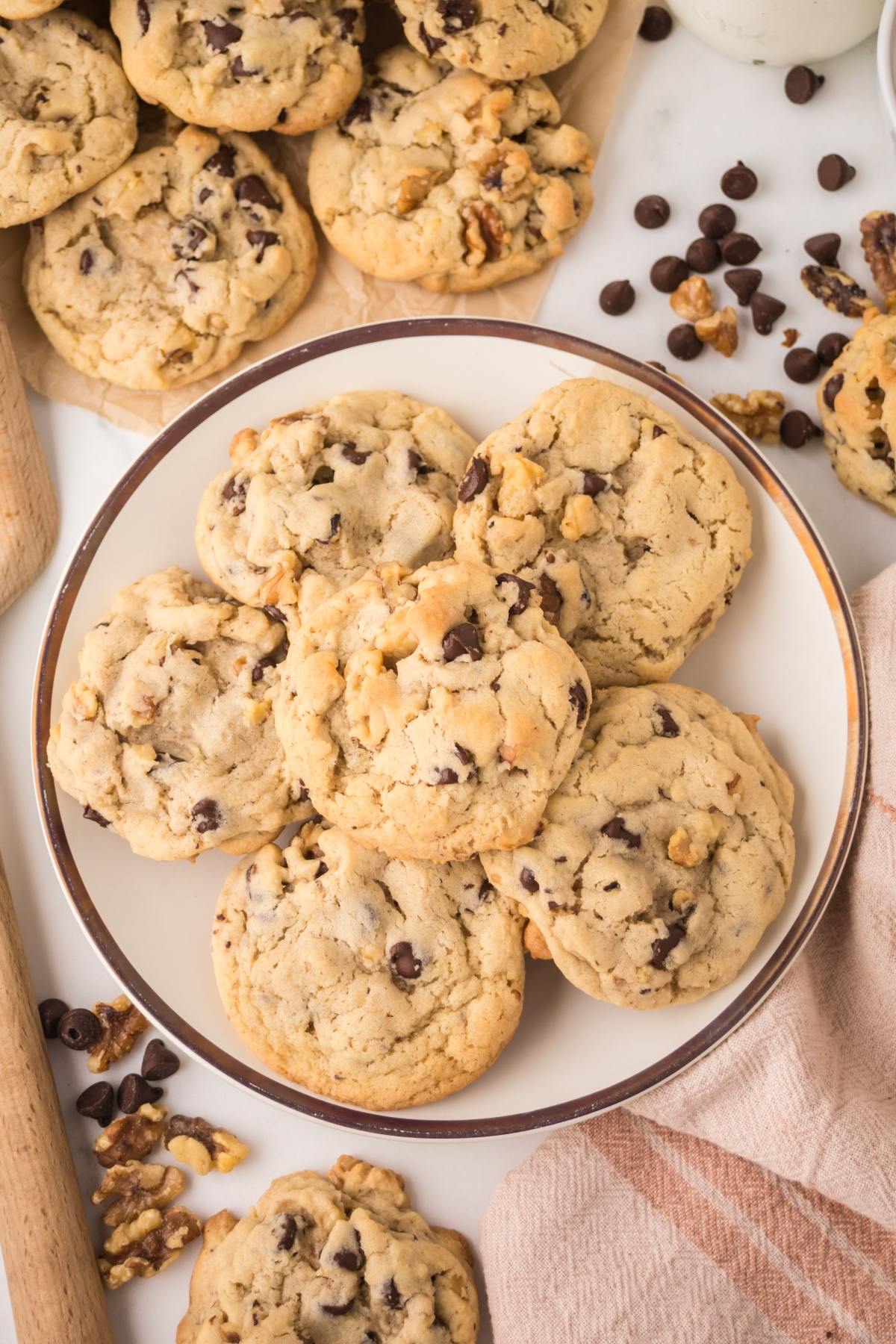 A plate of homemade chocolate chip cookies with walnuts, surrounded by more cookies and baking ingredients.