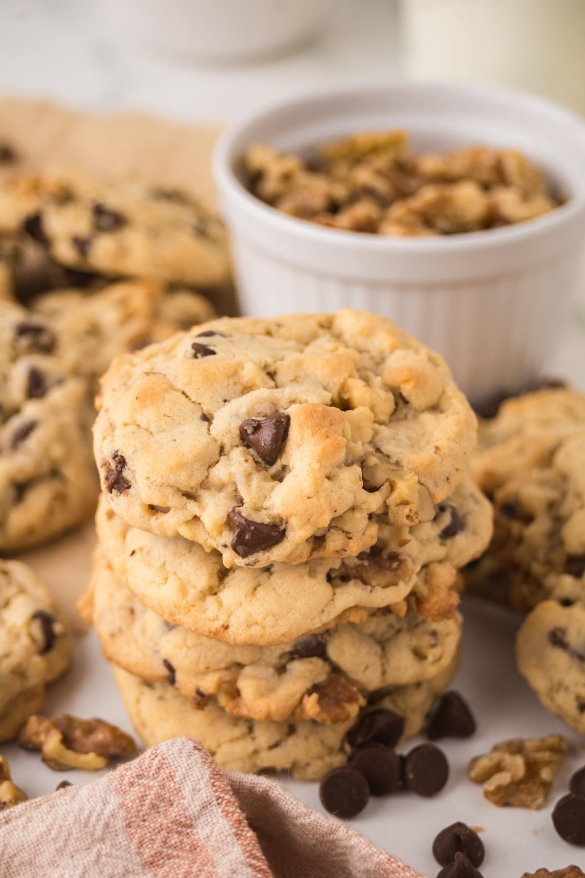 A stack of chocolate chip cookies with walnuts, surrounded by scattered chips and a bowl of chopped nuts.