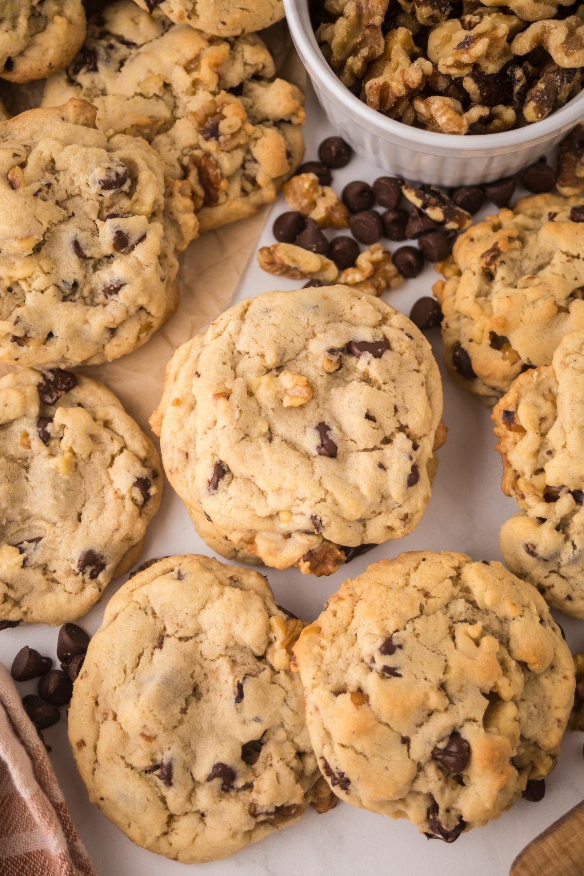 Chocolate chip cookies with walnuts are arranged next to a bowl of walnuts and scattered chocolate chips.