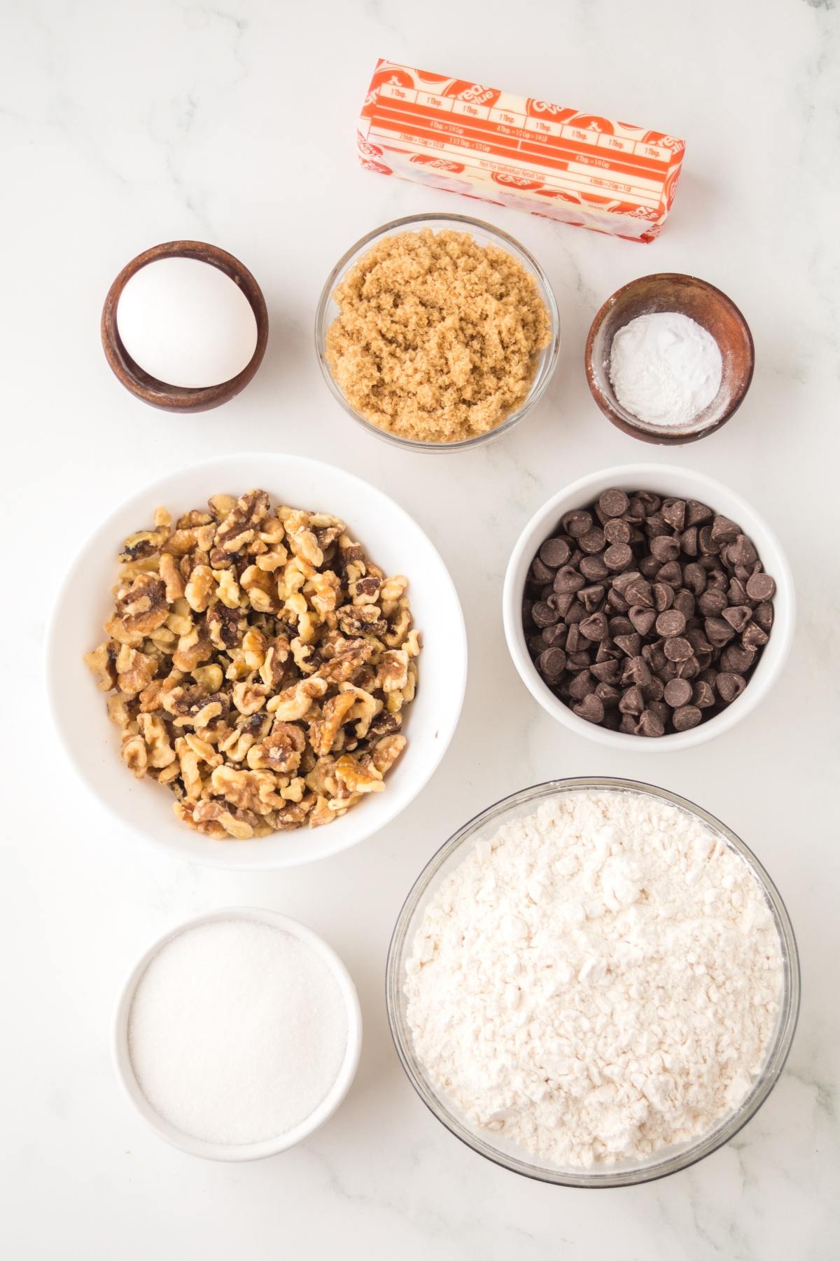Ingredients for baking: walnuts, flour, sugar, brown sugar, chocolate chips, butter, salt, and baking soda on a counter.