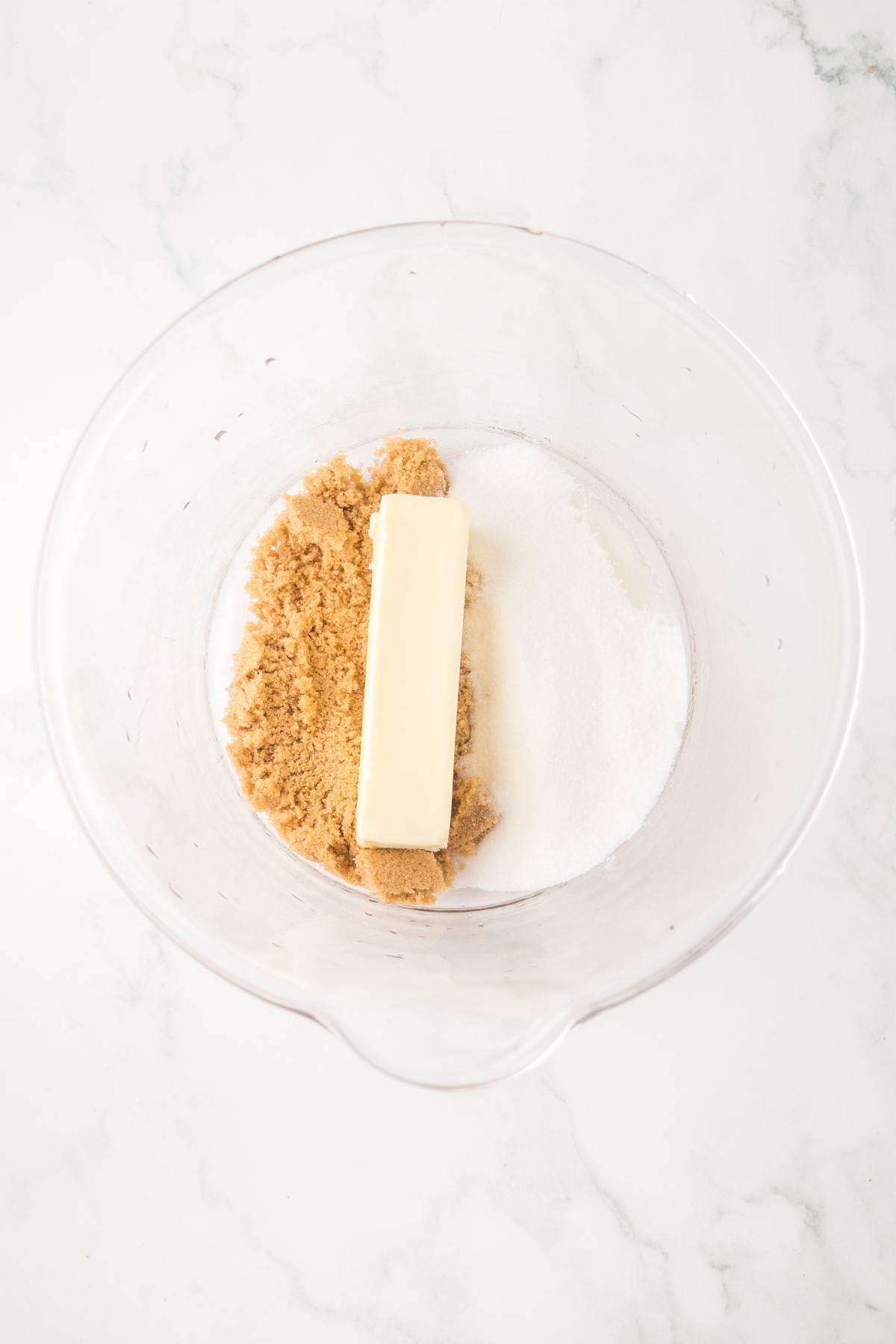 A bowl with a stick of butter, brown sugar, and white sugar on a white marble surface.
