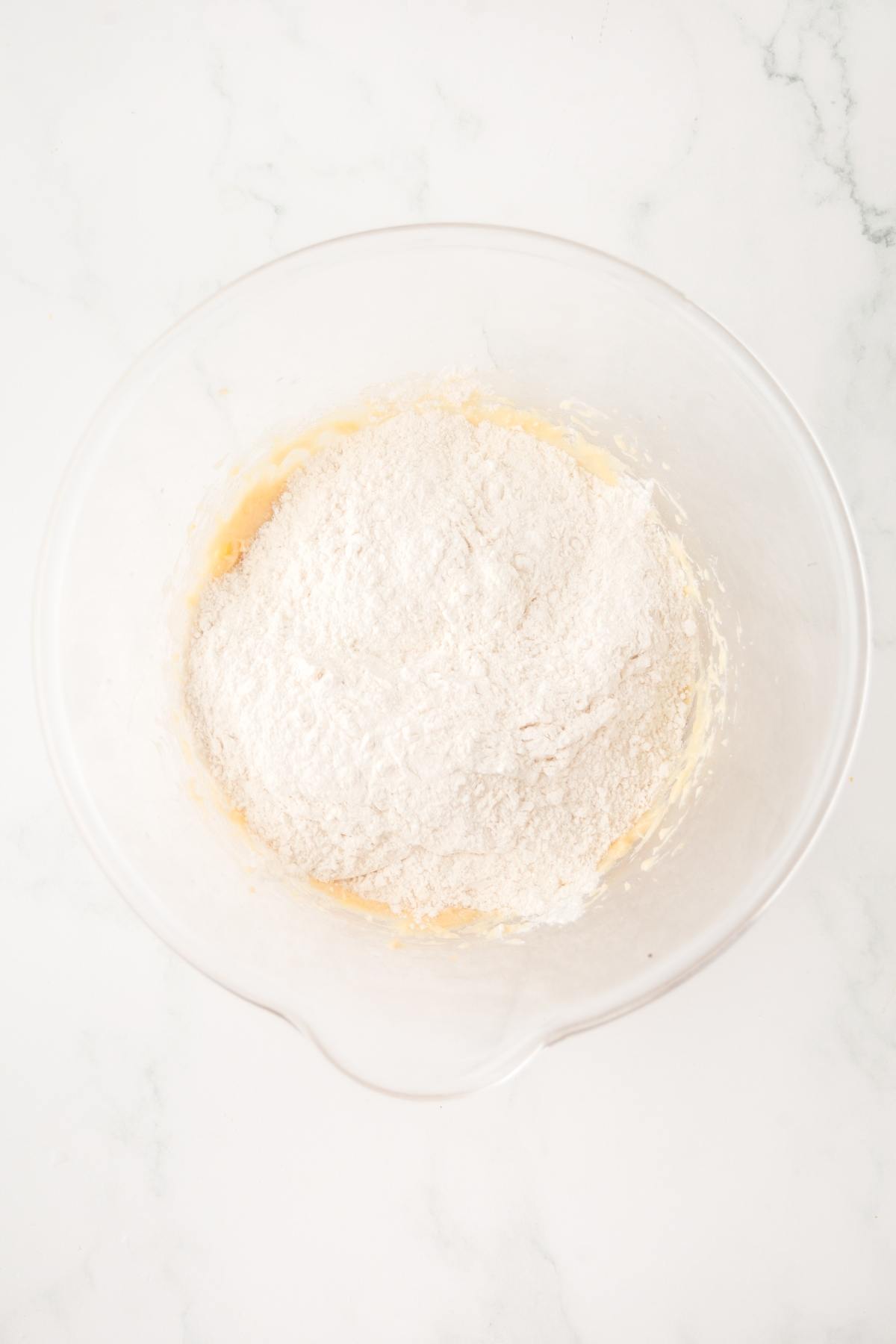 Clear mixing bowl with flour on top of a creamy batter, sitting on a white marble surface.