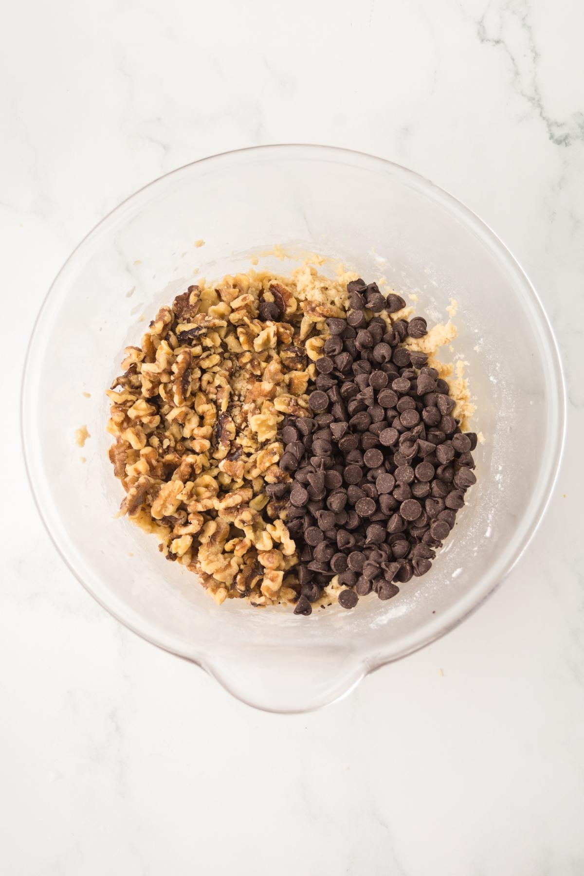 A glass bowl with cookie dough, chocolate chips, and chopped walnuts on a white marble surface.
