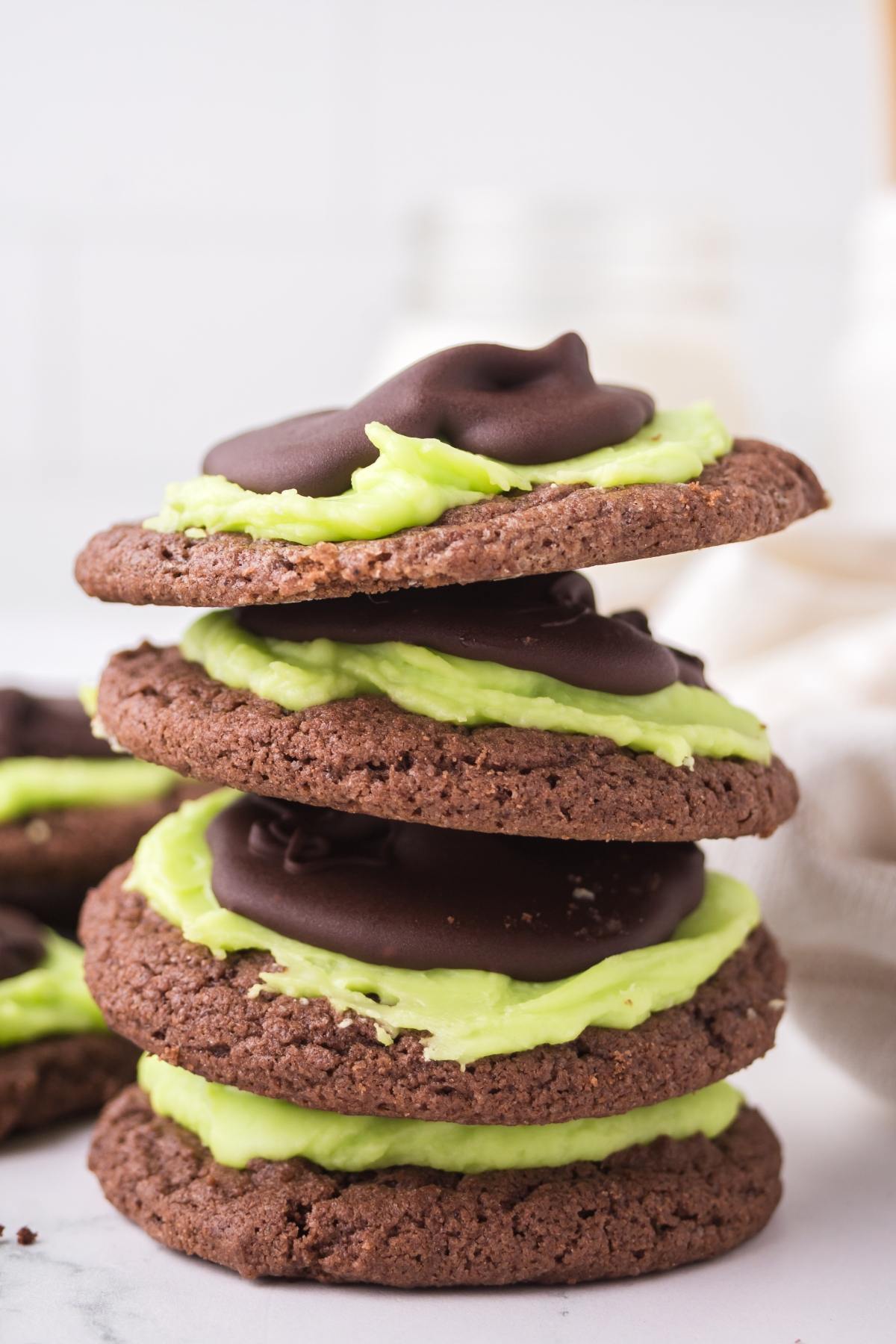 Three stacked chocolate cookies with green frosting and a chocolate topping, with more cookies in the background.