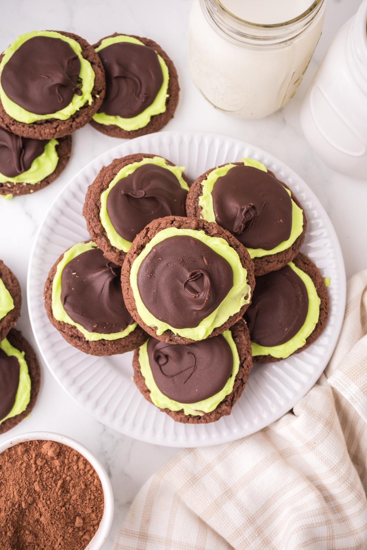 Plate of chocolate cookies with green frosting and chocolate topping, next to jars of milk on a white surface.
