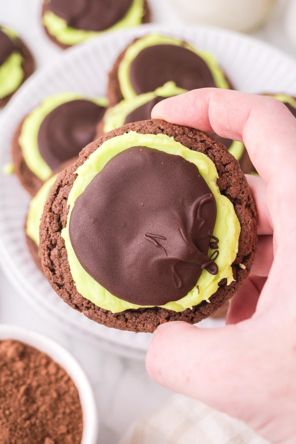 A hand holds a chocolate cookie topped with green frosting and a layer of chocolate.