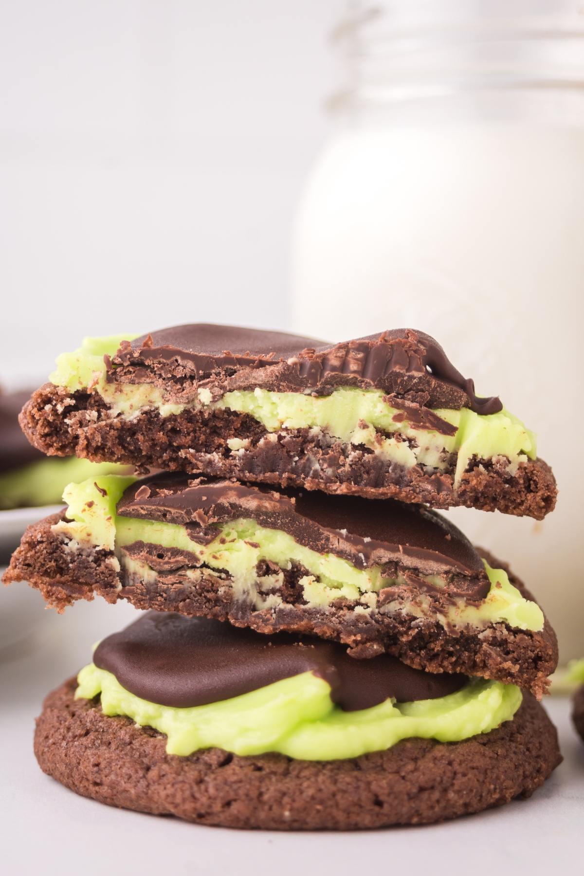 A chocolate cookie topped with green mint frosting and chocolate, cut in half to show the filling, in front of milk.