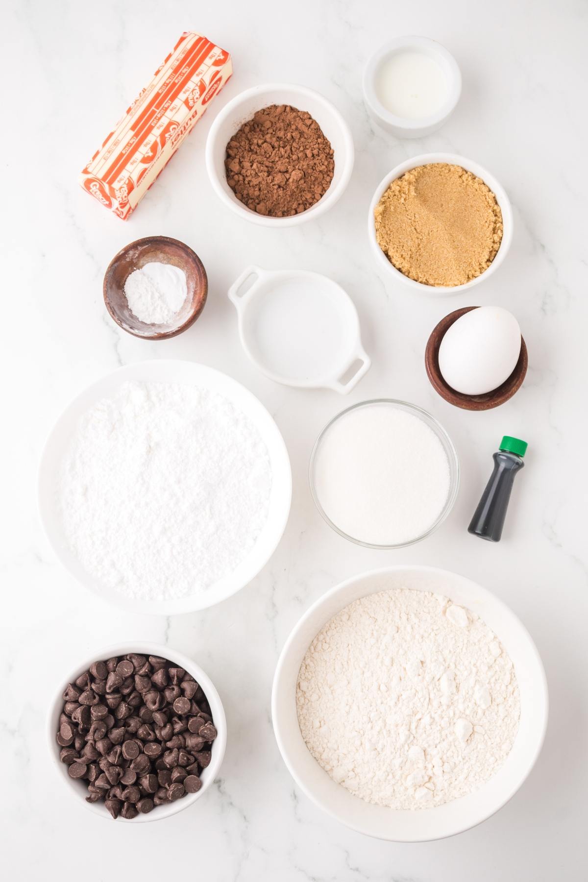 Baking ingredients arranged on a white surface, including flour, sugar, cocoa, egg, butter, and chocolate chips.