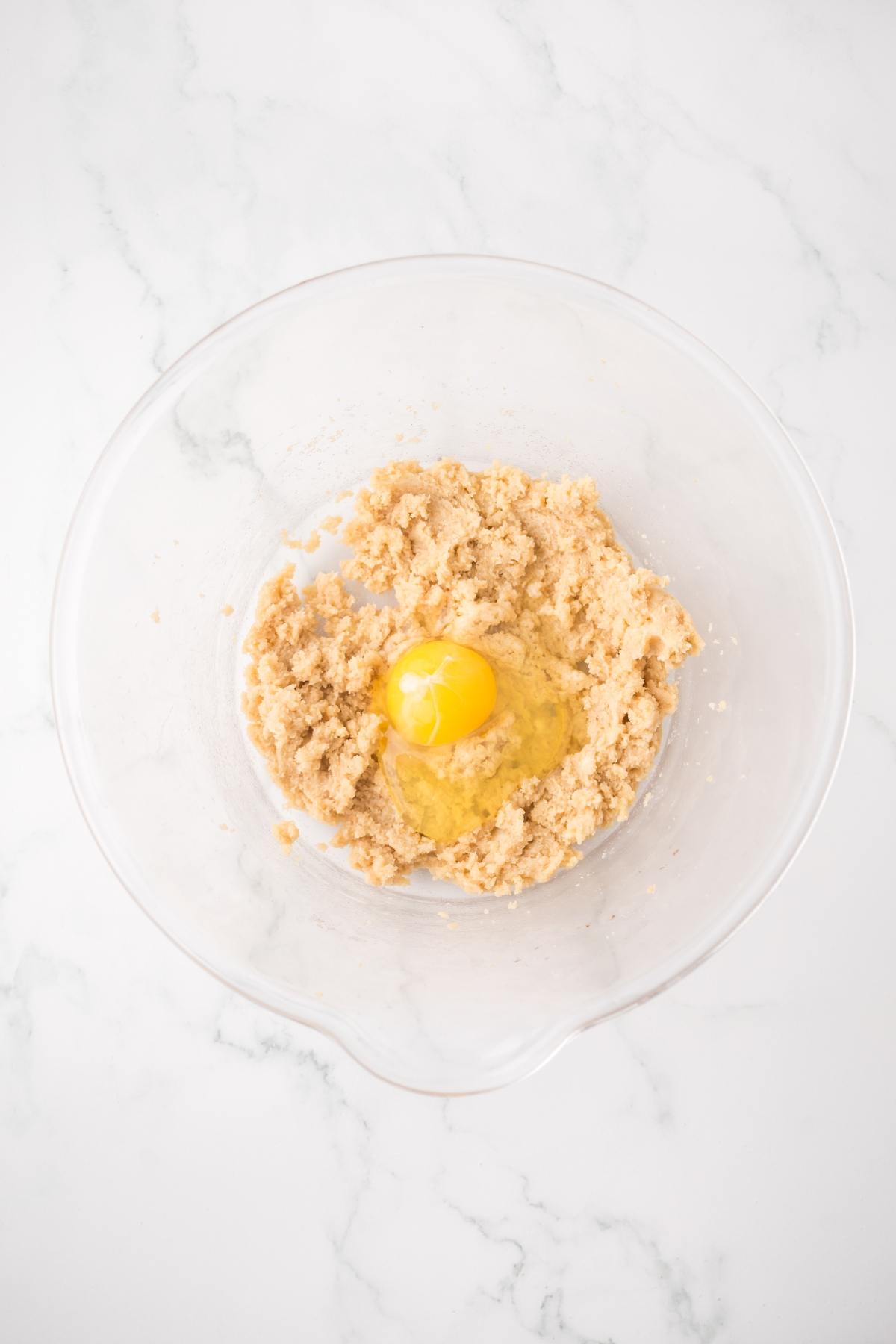 A mixing bowl with creamed sugar and butter, and a raw egg in the center, on a white marble surface.
