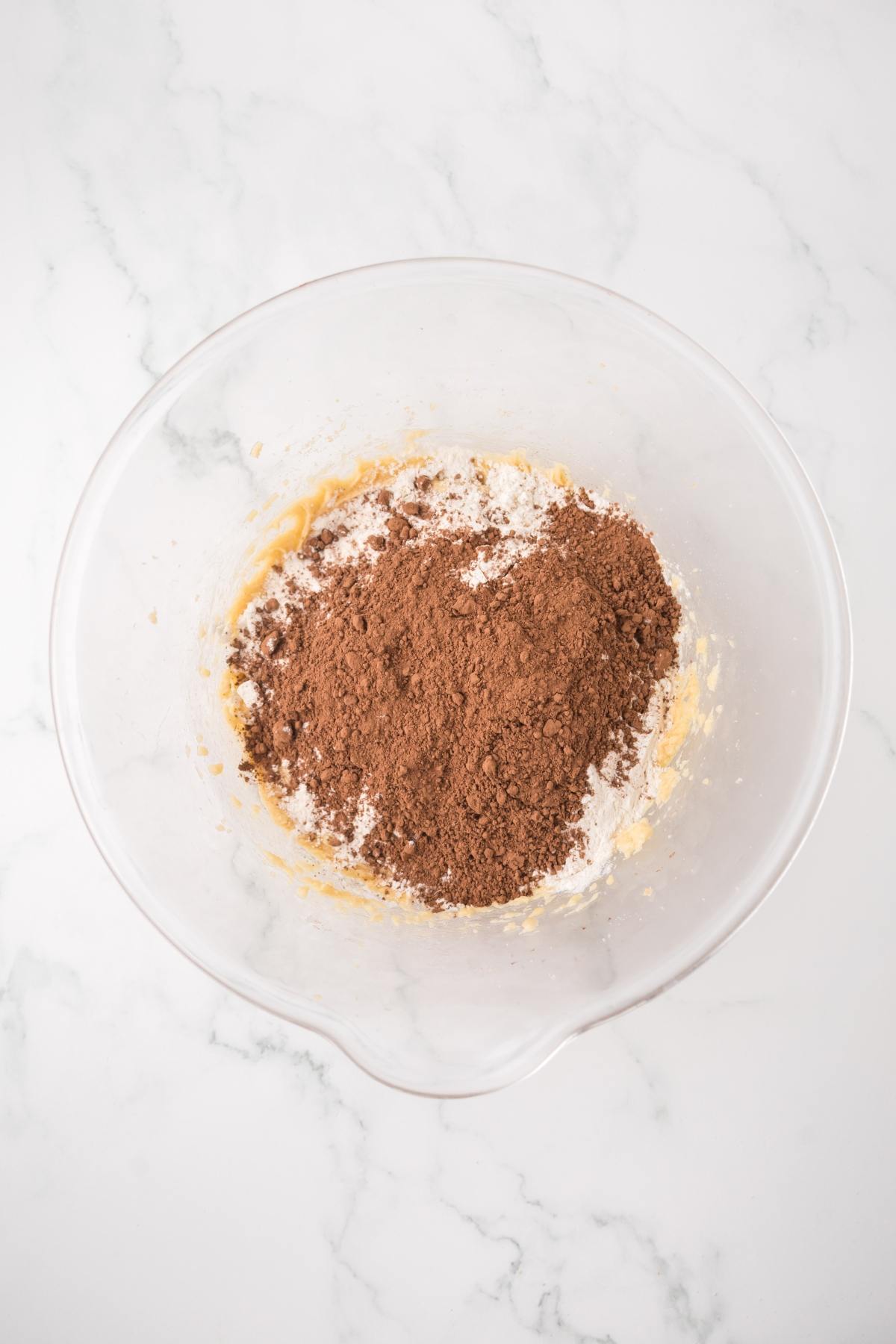 Clear glass mixing bowl with flour and cocoa powder on top of a light batter, on a white marble surface.