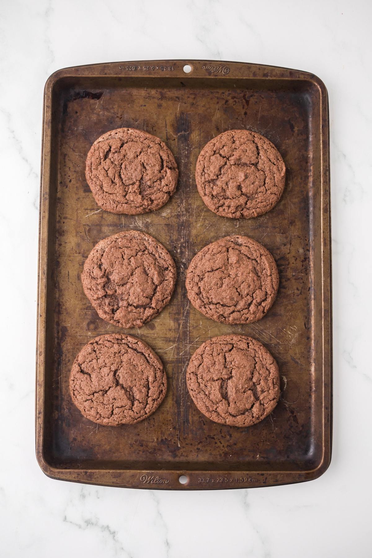 Six chocolate cookies on a baking sheet, arranged in two columns of three, on a white marble surface.