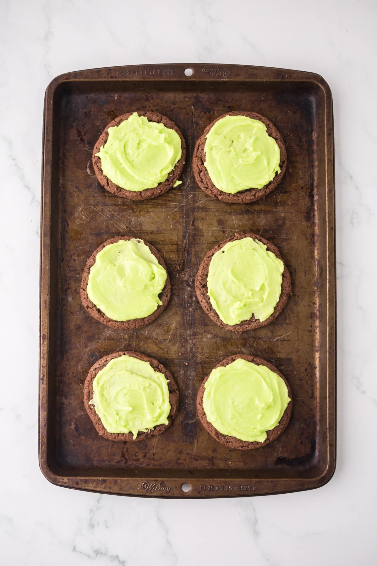 Six chocolate cookies with bright green frosting on a baking sheet, viewed from above.