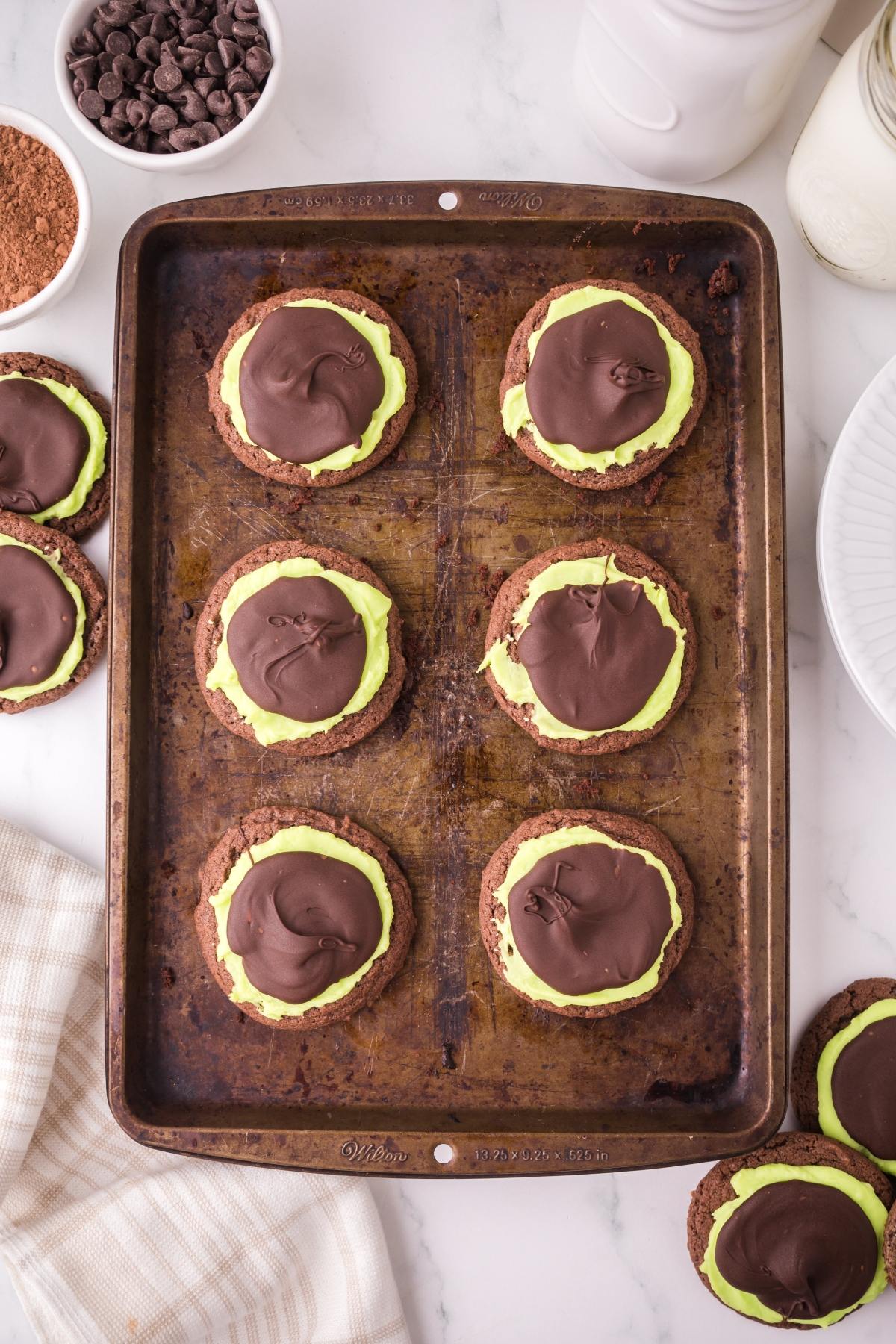 Six chocolate cookies with green frosting and chocolate topping on a baking sheet, surrounded by baking items.