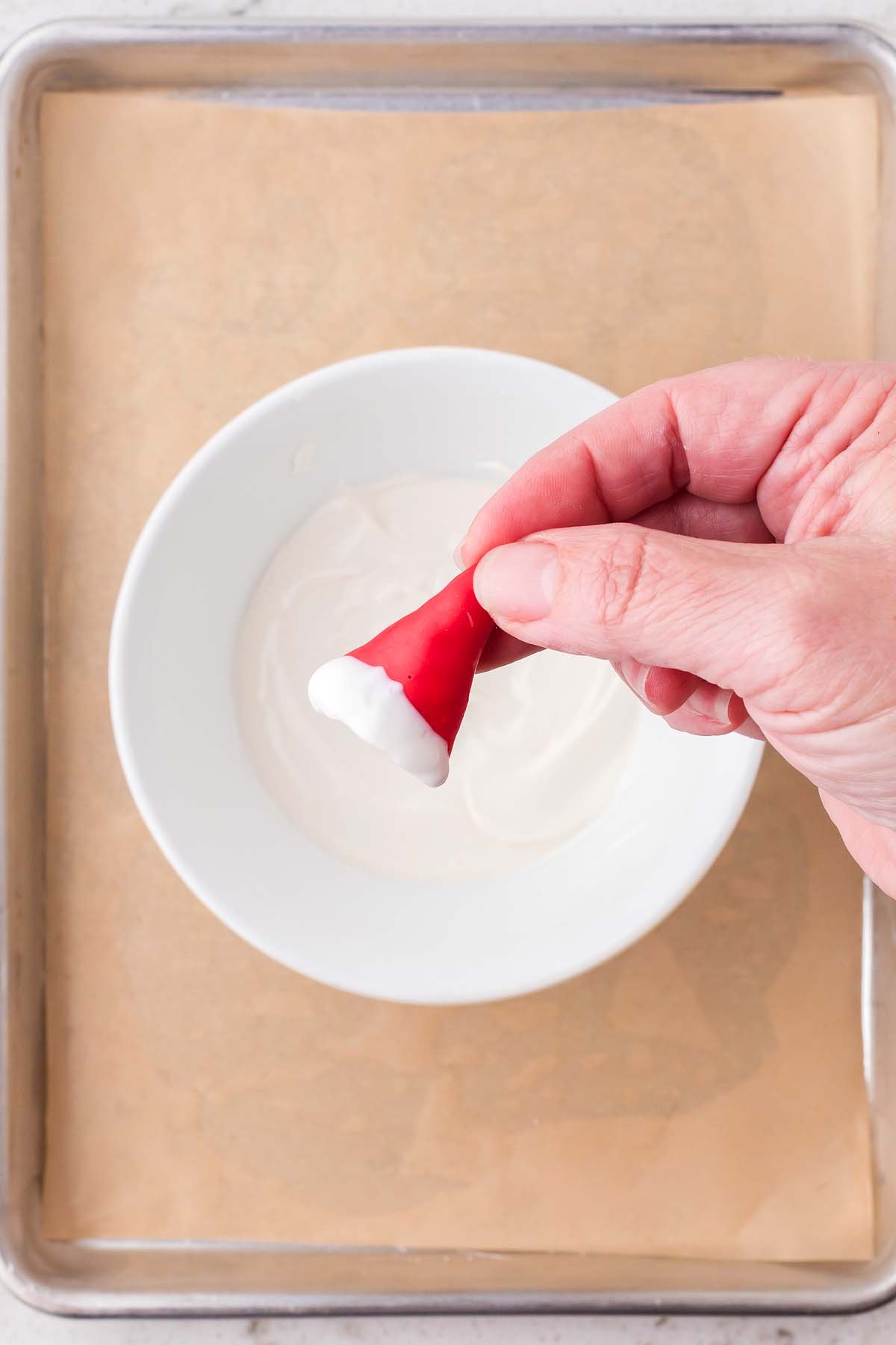 Hand holding a red candy dipped in white frosting over a bowl, resembling a Santa hat—perfect for festive treats like Christmas Bugles.