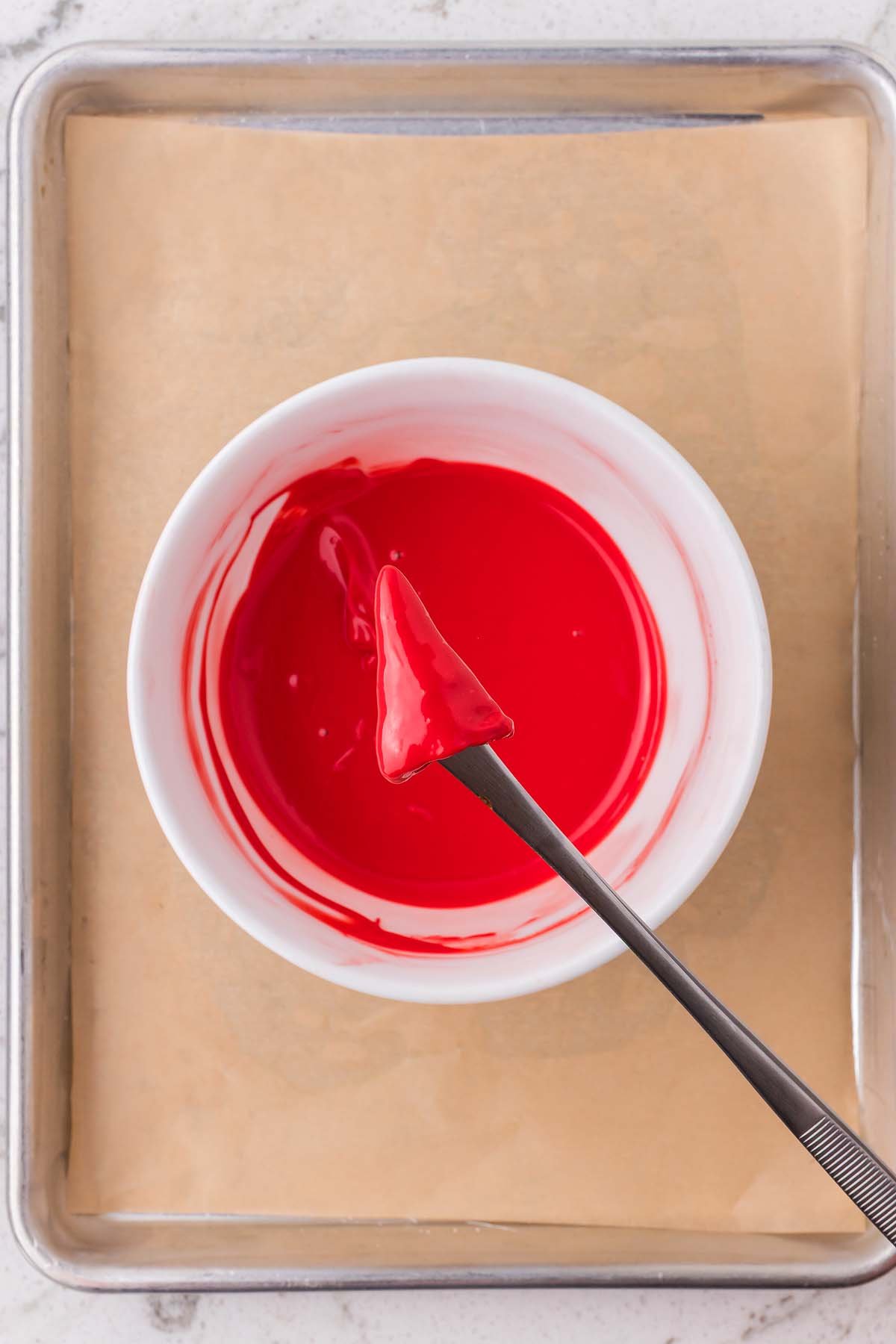 A fork holds a red candy-coated strawberry over a bowl of red melted coating on a baking sheet, perfect for pairing with festive Christmas Bugles treats.