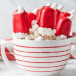 Christmas Cakesicles decorated as Santa hats are served in a striped mug filled with marshmallows, with Christmas trees in the background.