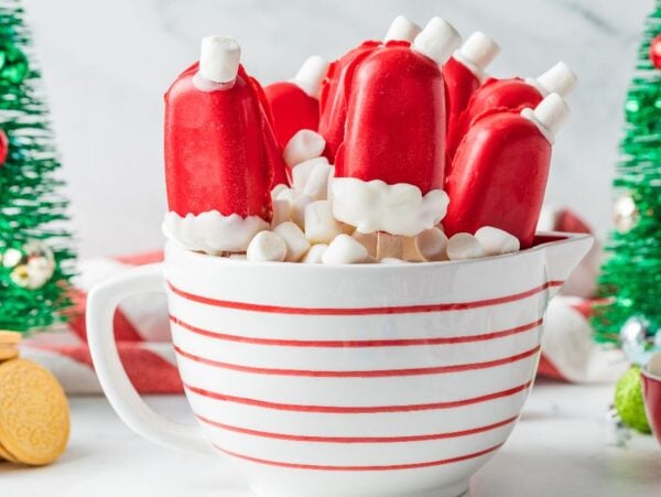 Christmas Cakesicles decorated as Santa hats are served in a striped mug filled with marshmallows, with Christmas trees in the background.