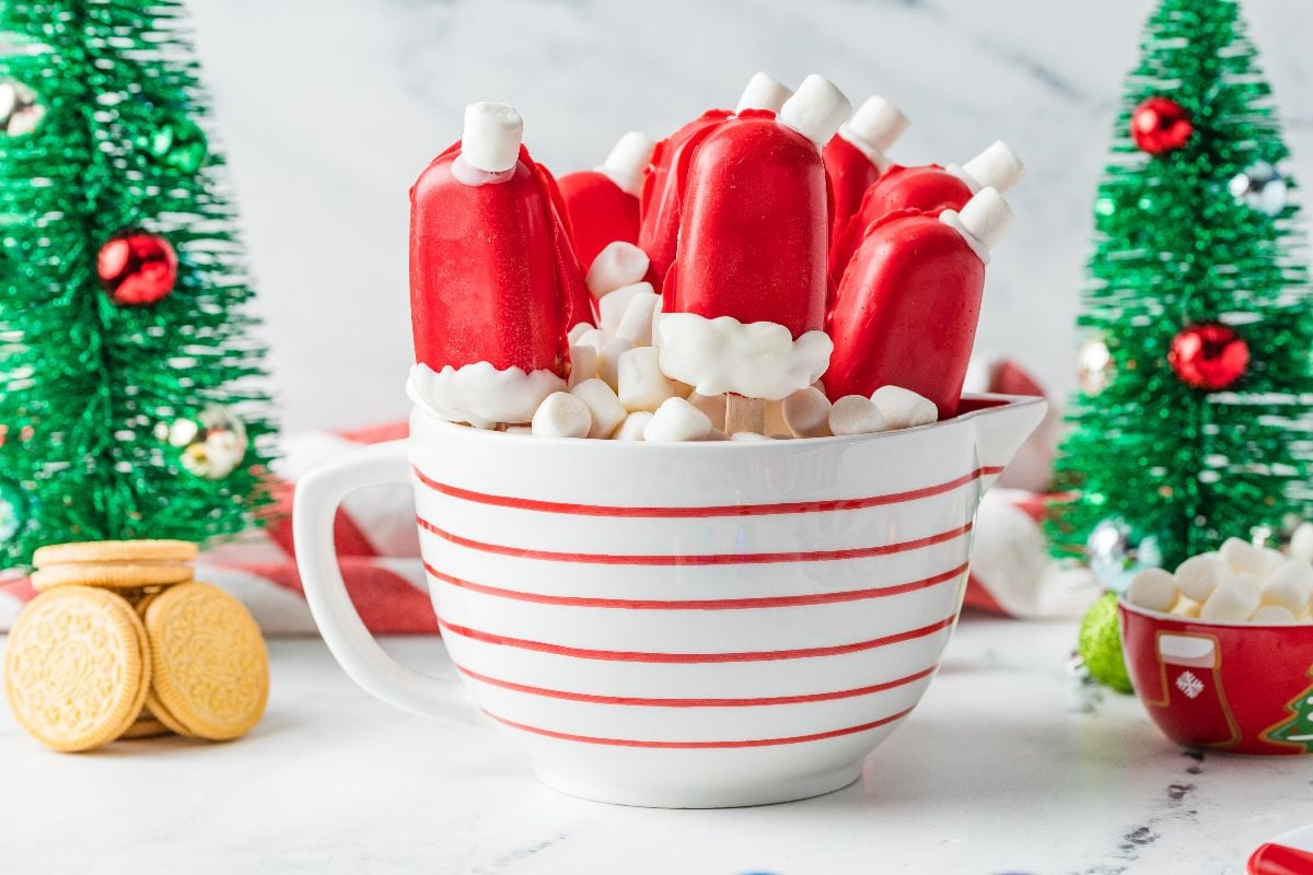 Santa hat-shaped Christmas Cakesicles on sticks in a striped mug with mini marshmallows, surrounded by festive decorations.