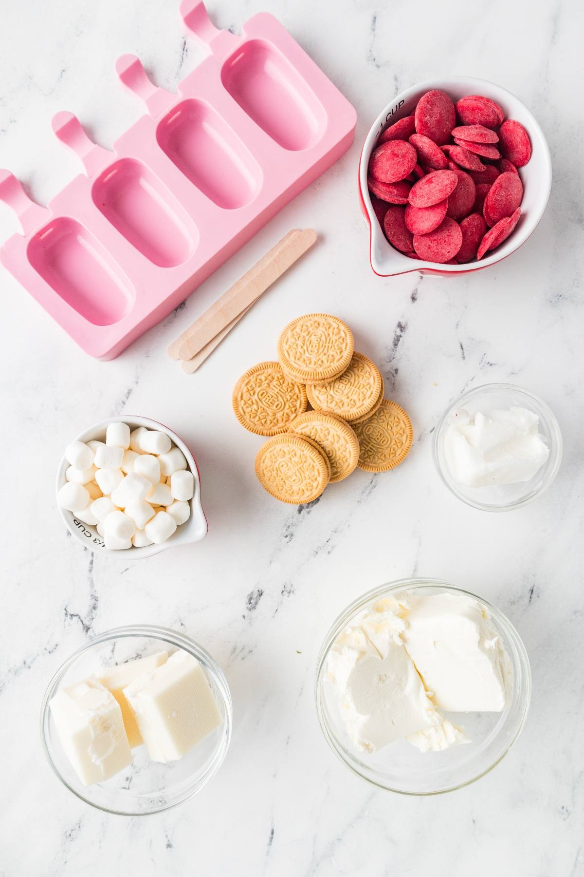 Ice cream pop molds, cookies, marshmallows, pink wafers, and dairy ingredients on a marble surface.