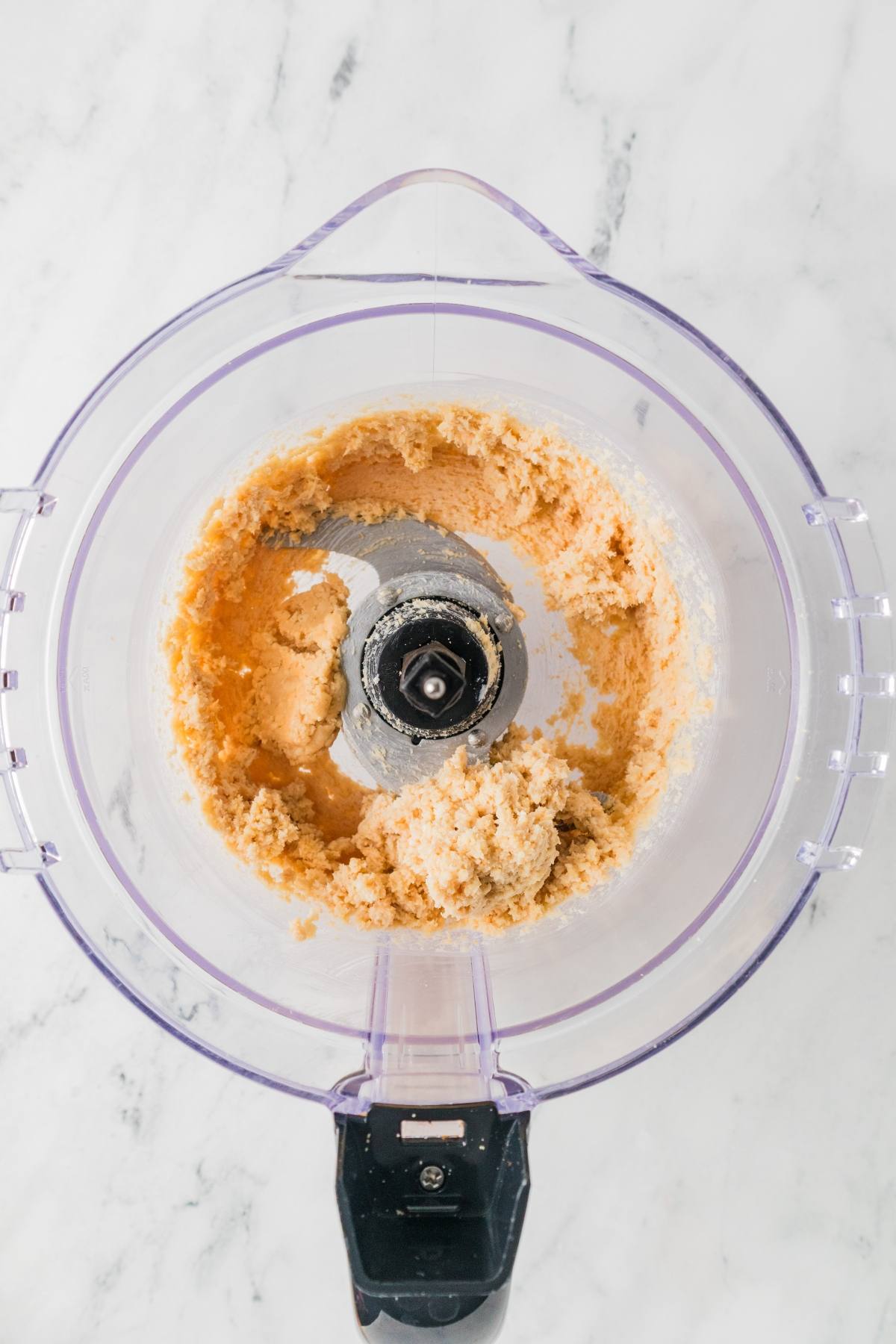 A food processor bowl with creamy dough mixture inside, viewed from above on a white surface.