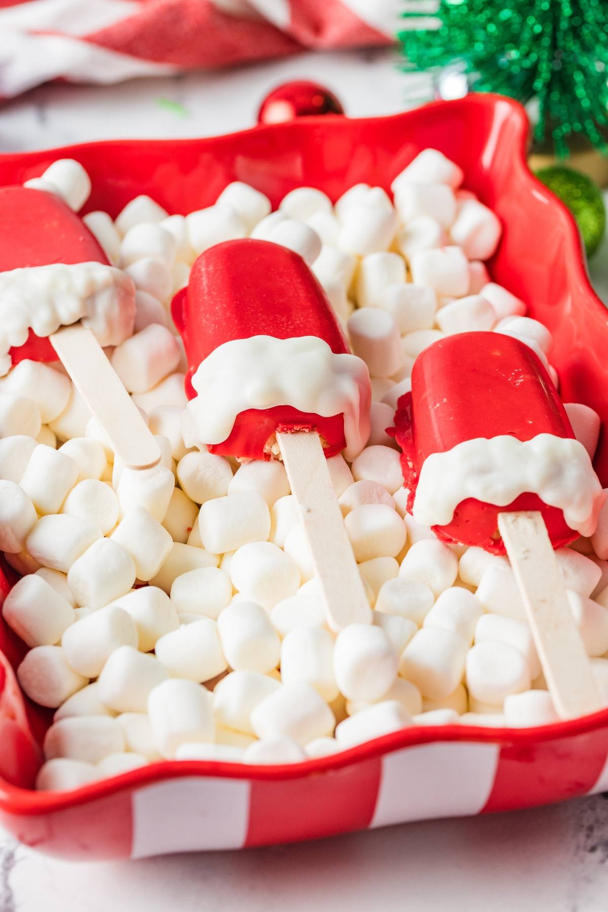 Three red popsicles with white chocolate trim on sticks, set on mini marshmallows in a red dish.