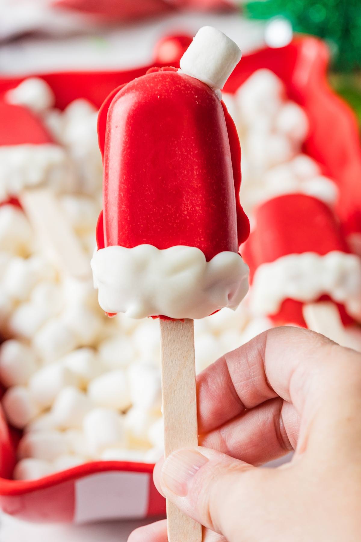 A hand holds a Santa hat-shaped treat on a stick, decorated with red icing and white marshmallows.