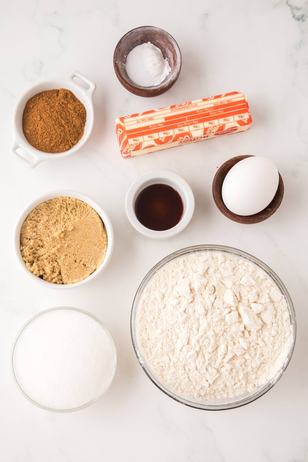 Baking ingredients on a white surface: flour, sugars, butter, egg, vanilla, baking soda, and cinnamon.