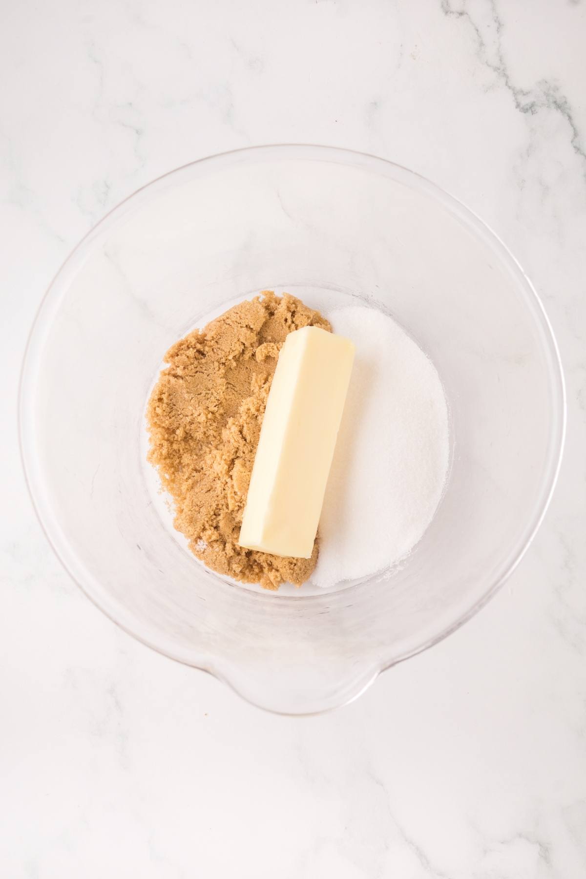 A glass bowl with a stick of butter, brown sugar, and white sugar on a white marble surface.