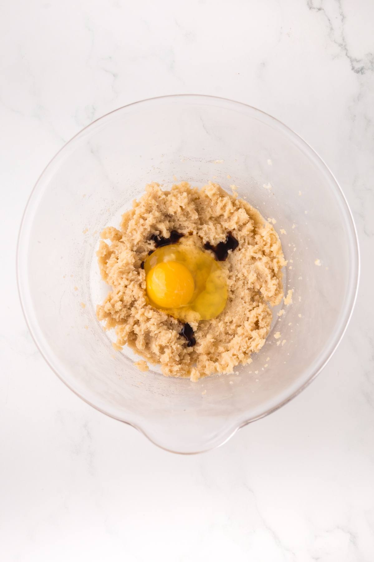 A glass bowl with cookie dough, an egg, and vanilla extract on a white surface.