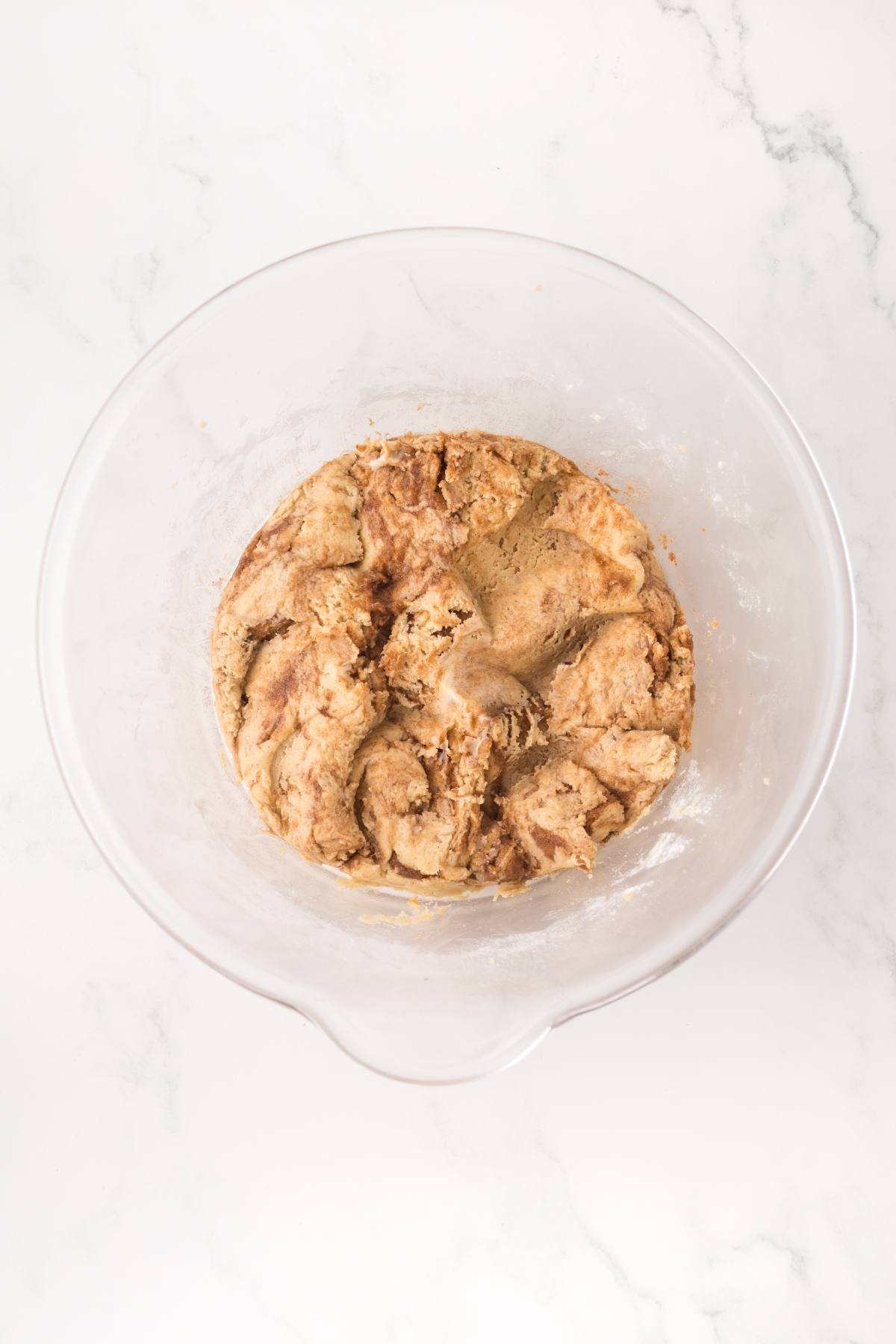 A glass bowl with swirled cinnamon roll dough on a white marble surface.