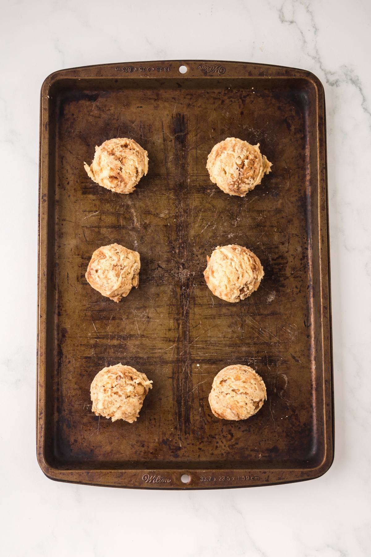 Six unbaked biscuit dough balls on a baking sheet, spaced evenly apart on a light surface.