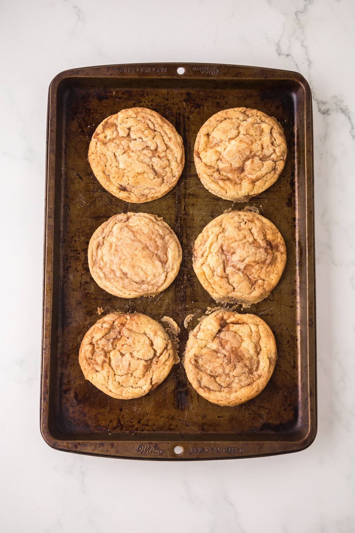 Six large cookies on a baking sheet, placed on a white marble surface.