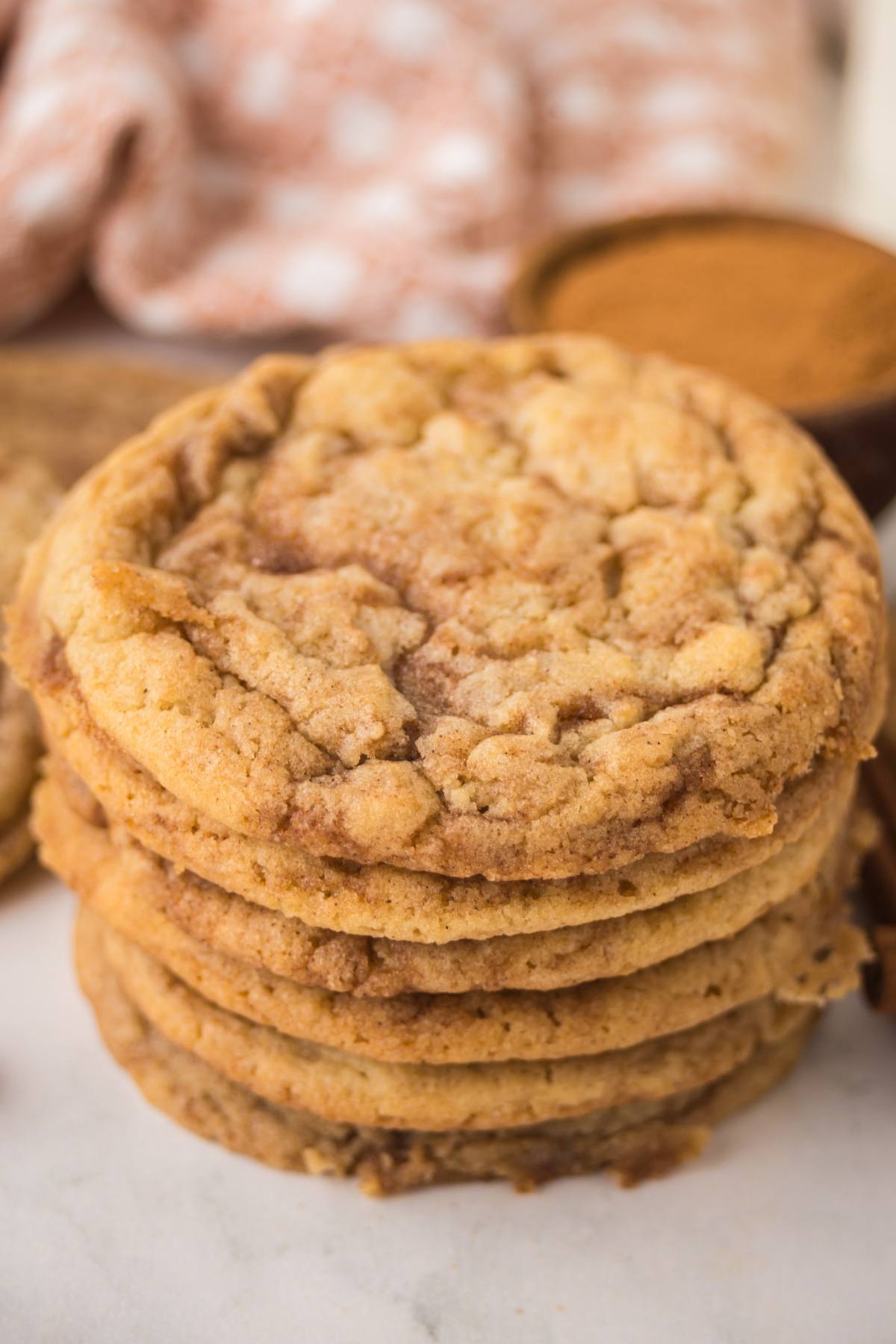 A stack of chewy cinnamon roll cookies on a white surface with cinnamon in the background.