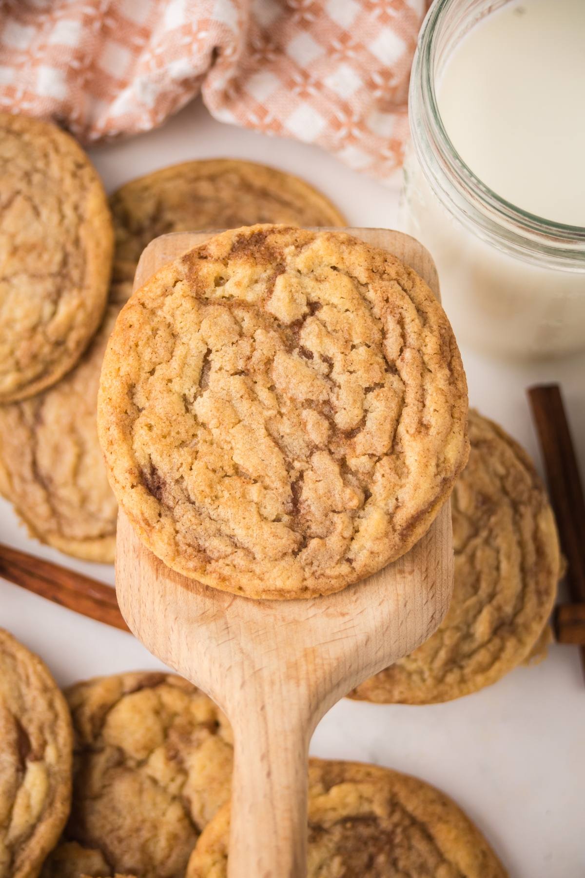 A wooden spatula holds a chewy cookie above more cookies, with a glass of milk and cinnamon sticks nearby.