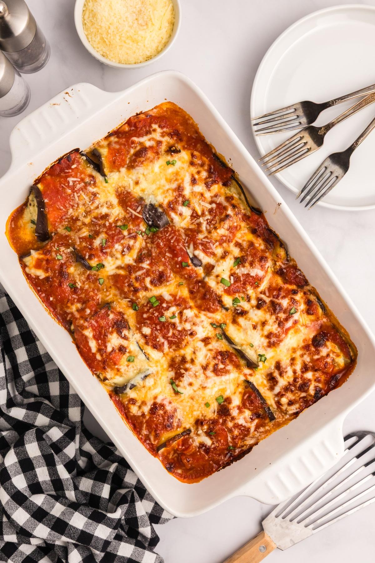 Baked cheesy eggplant rollatini in a white dish, with plates, forks, grated cheese, and a checkered napkin nearby.