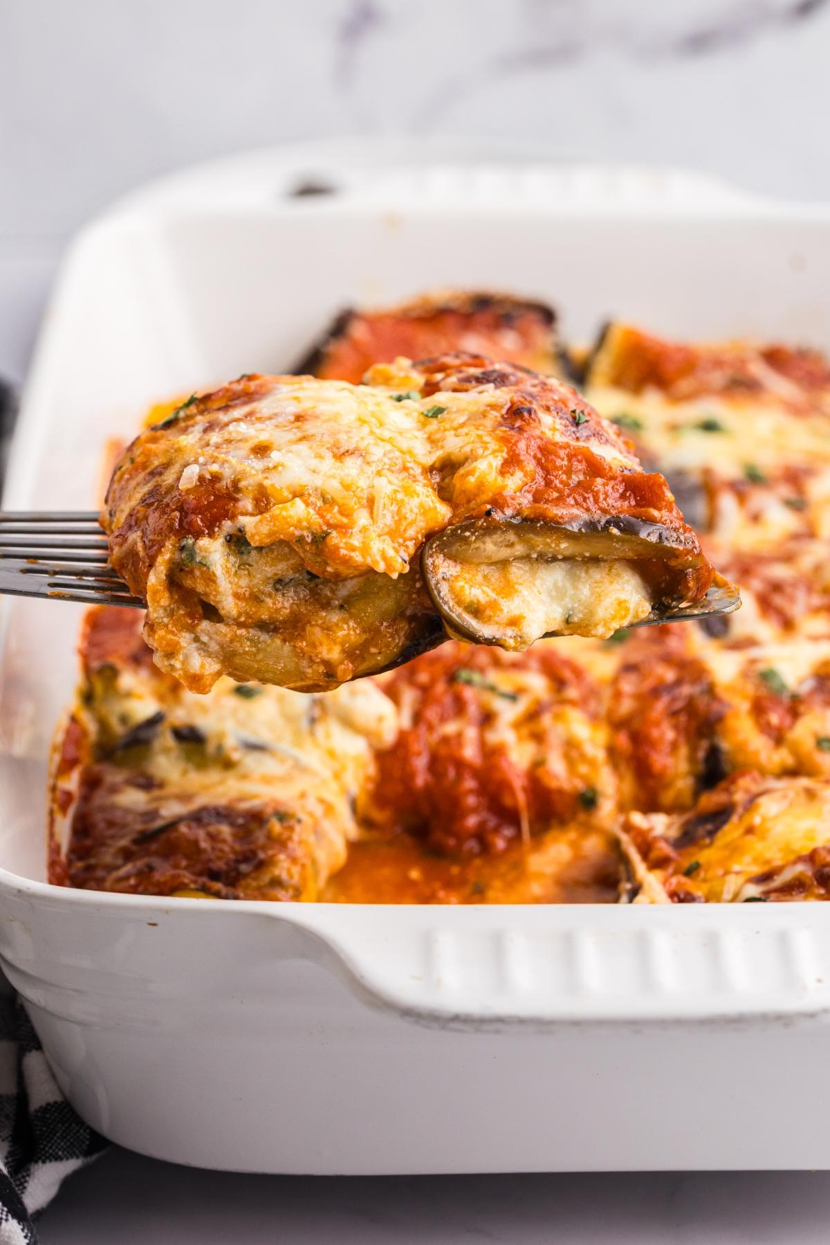 A fork lifts a cheesy, baked eggplant roll from a casserole dish filled with sauce and melted cheese.