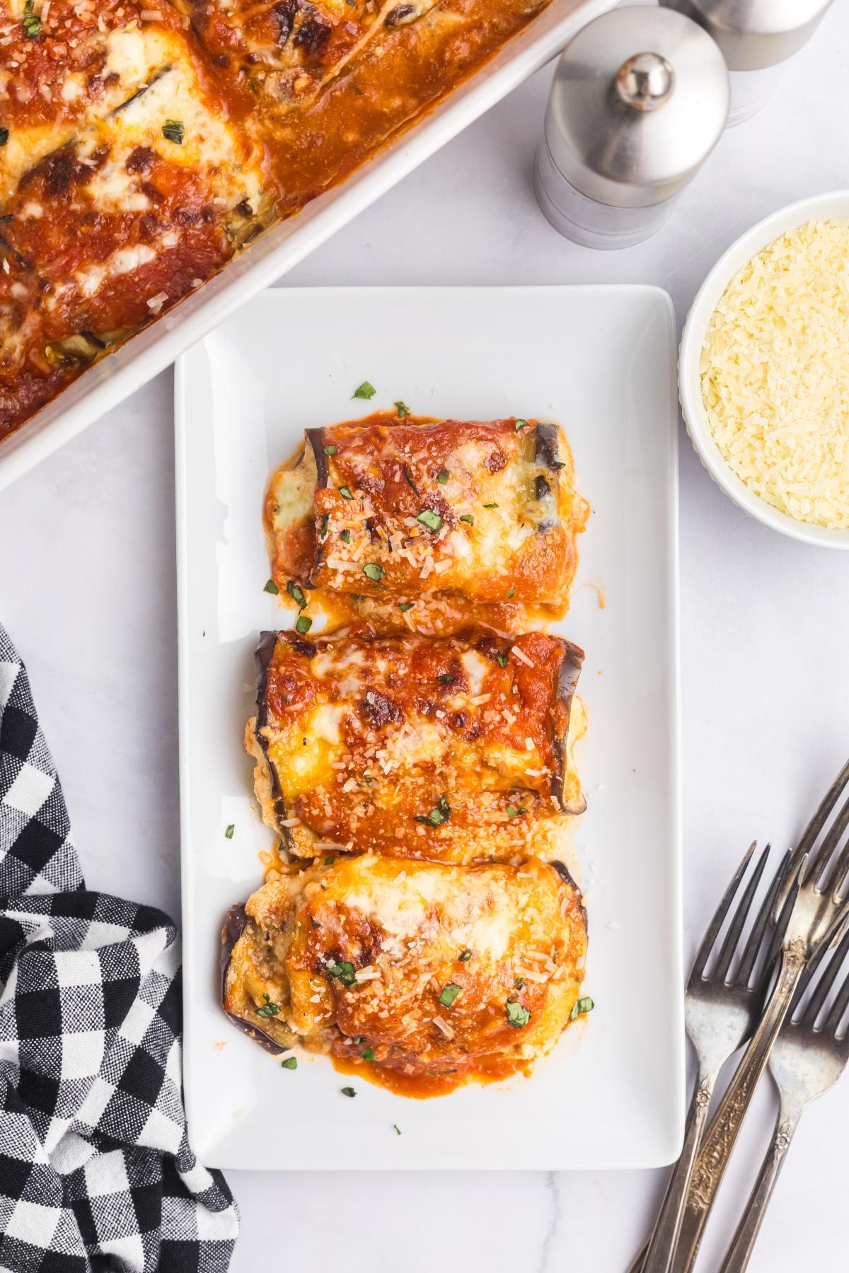 Three pieces of cheesy eggplant rollatini served on a white plate, with sauce, grated cheese, and utensils nearby.